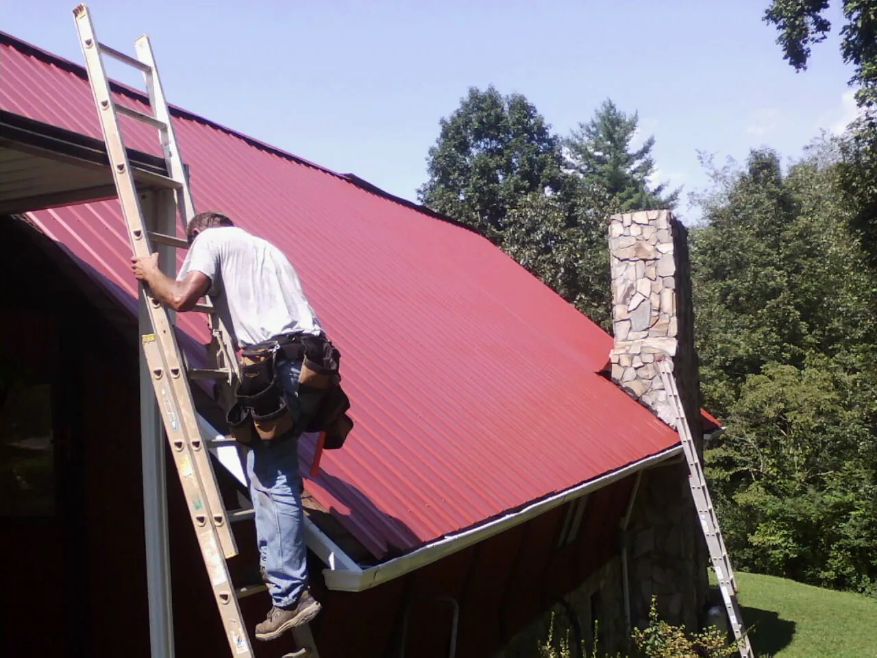 Roofer on a ladder beside a red metal roof and stone chimney — Soffit & Fascia Repair by Roofing Brothers in Fort Ransom